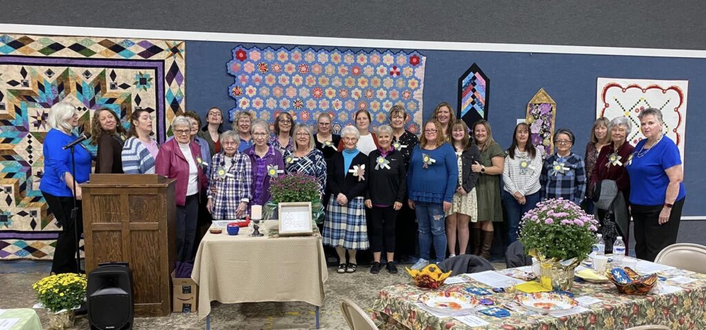 A women's tea event in the gym at Rosedale Friends Church, with many women standing in front of a wall of quilts